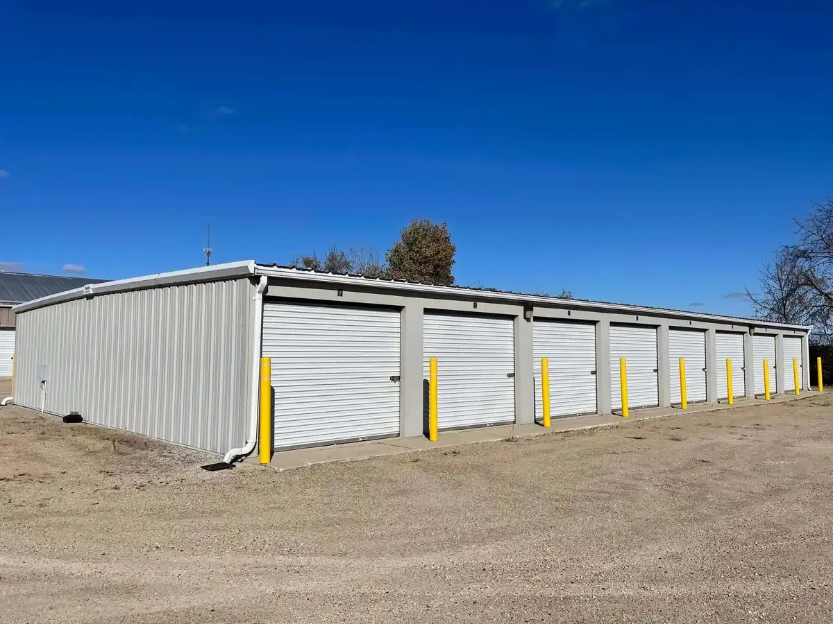 Long metal storage building with multiple roll-up doors, yellow safety posts, and a gravel lot under a clear blue sky.