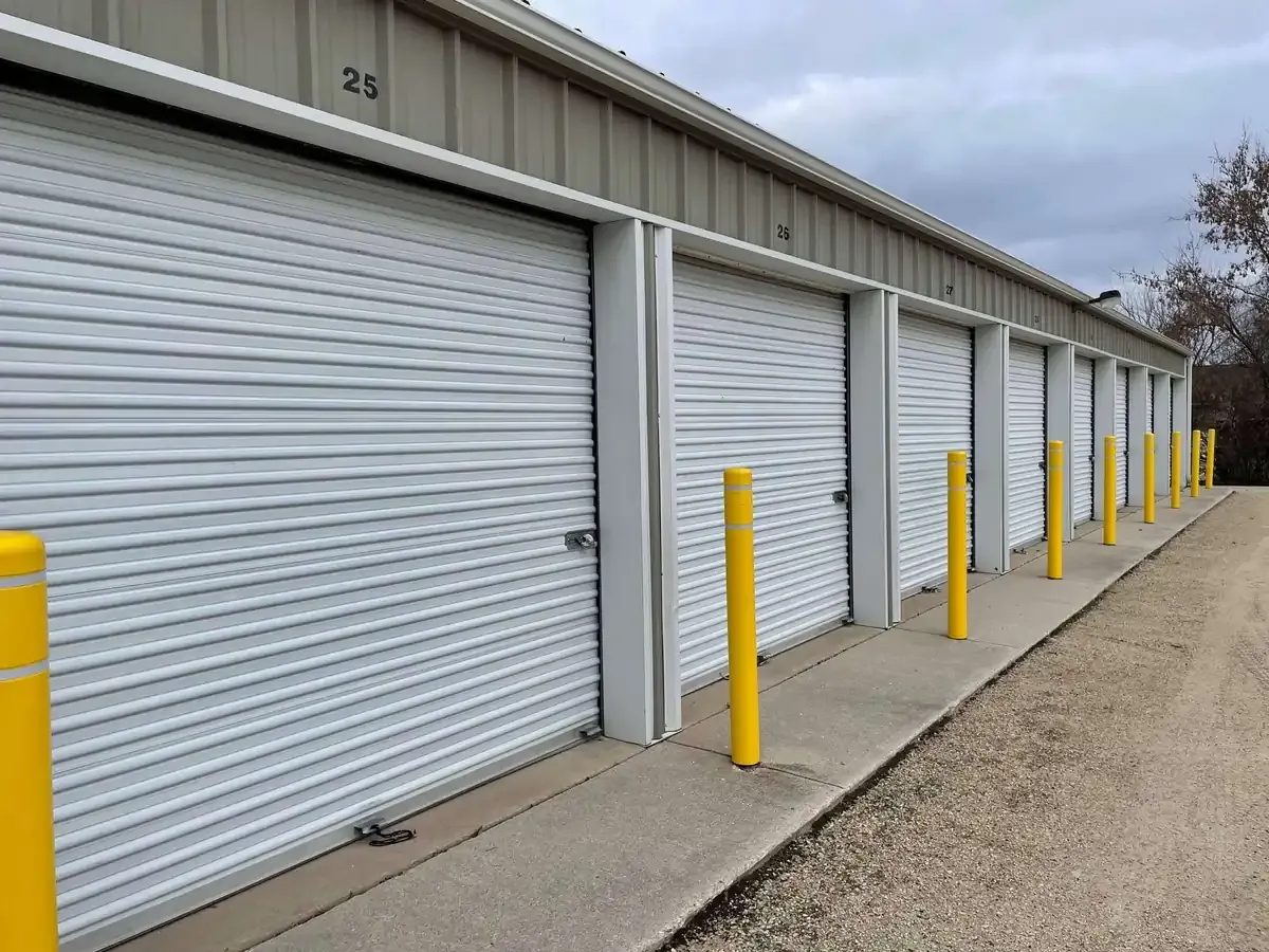 Storage units with white roll-up doors, numbered 25 to 27, separated by gray metal frames, with yellow protective...