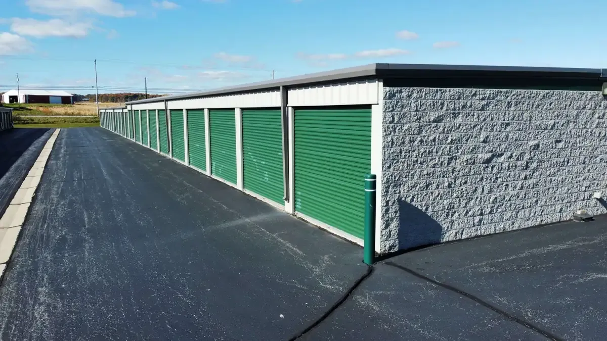 Storage units with green roll-up doors, outside parking area, and security fencing under a blue sky.