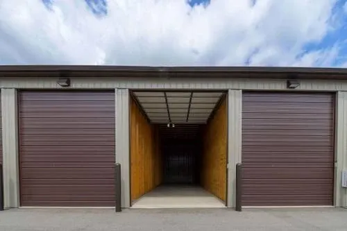 Open storage unit with wooden walls and metal roof, flanked by two closed roll-up garage doors, in a storage facility.