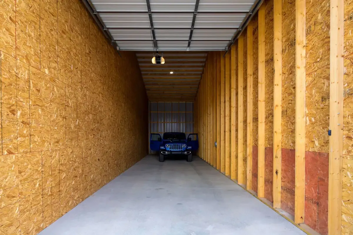 A blue Jeep parked inside a partially finished storage unit with plywood walls, an open garage door, and a concrete floor.