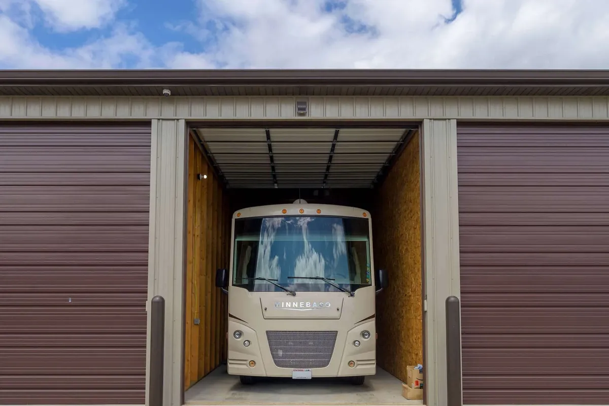Compact beige shuttle bus parked inside a partially open storage unit with brown metal roll-up doors and wooden interior...