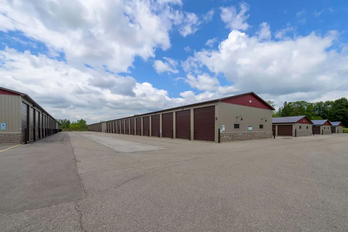 Long row of storage units with roll-up doors in a spacious, paved outdoor storage facility under a partly cloudy sky.