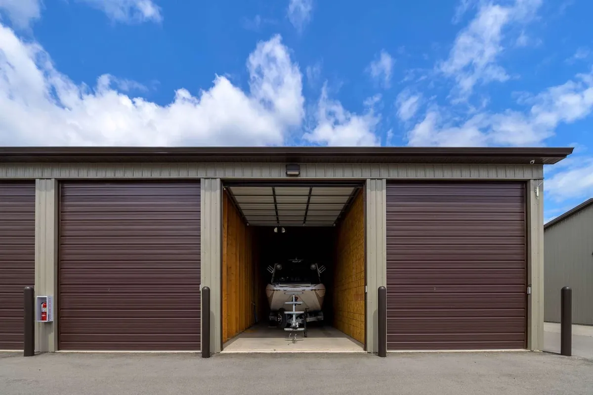Storage unit with a boat inside, surrounded by metal walls and roller doors, under a blue sky with clouds.