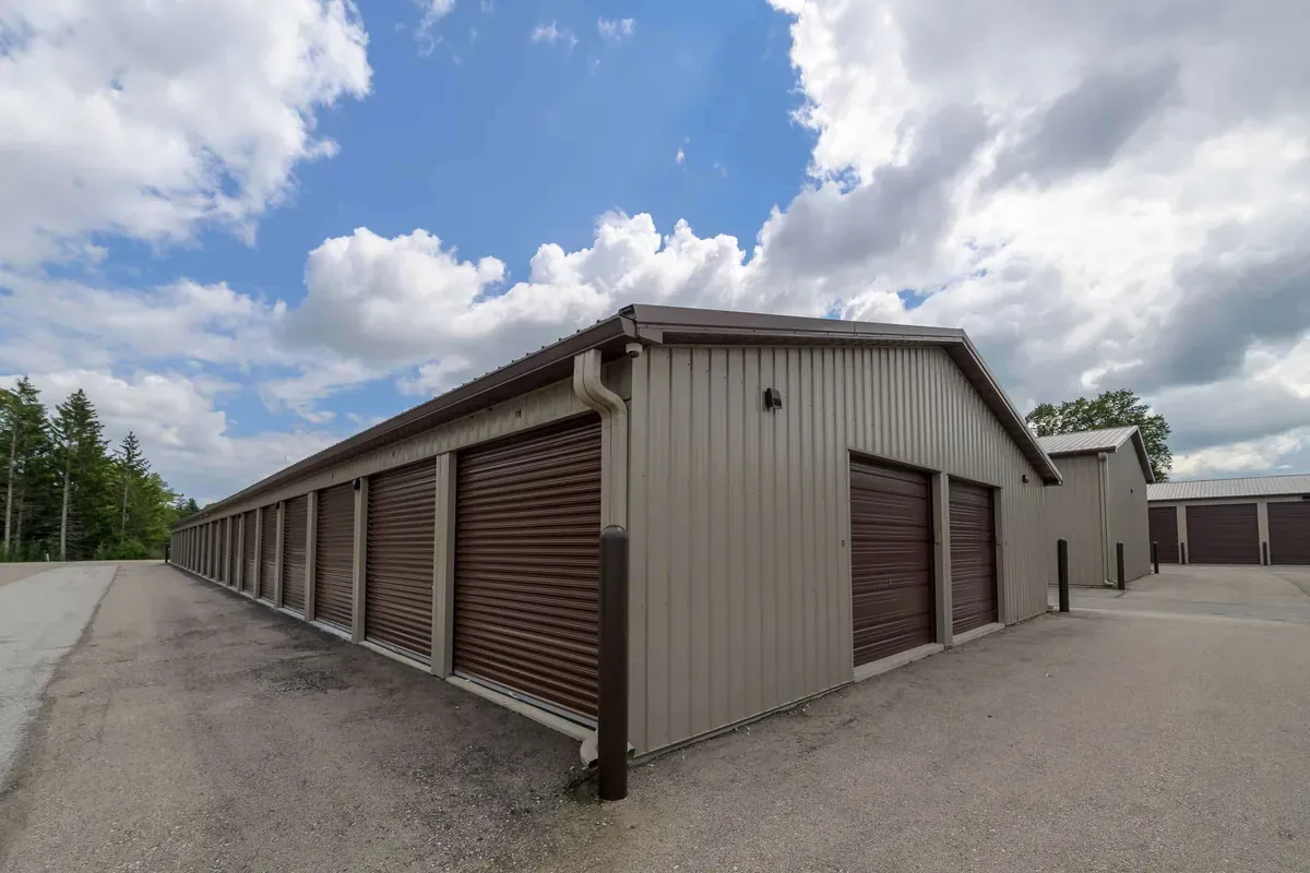 Long row of beige metal storage units with brown roll-up doors under a partly cloudy sky.