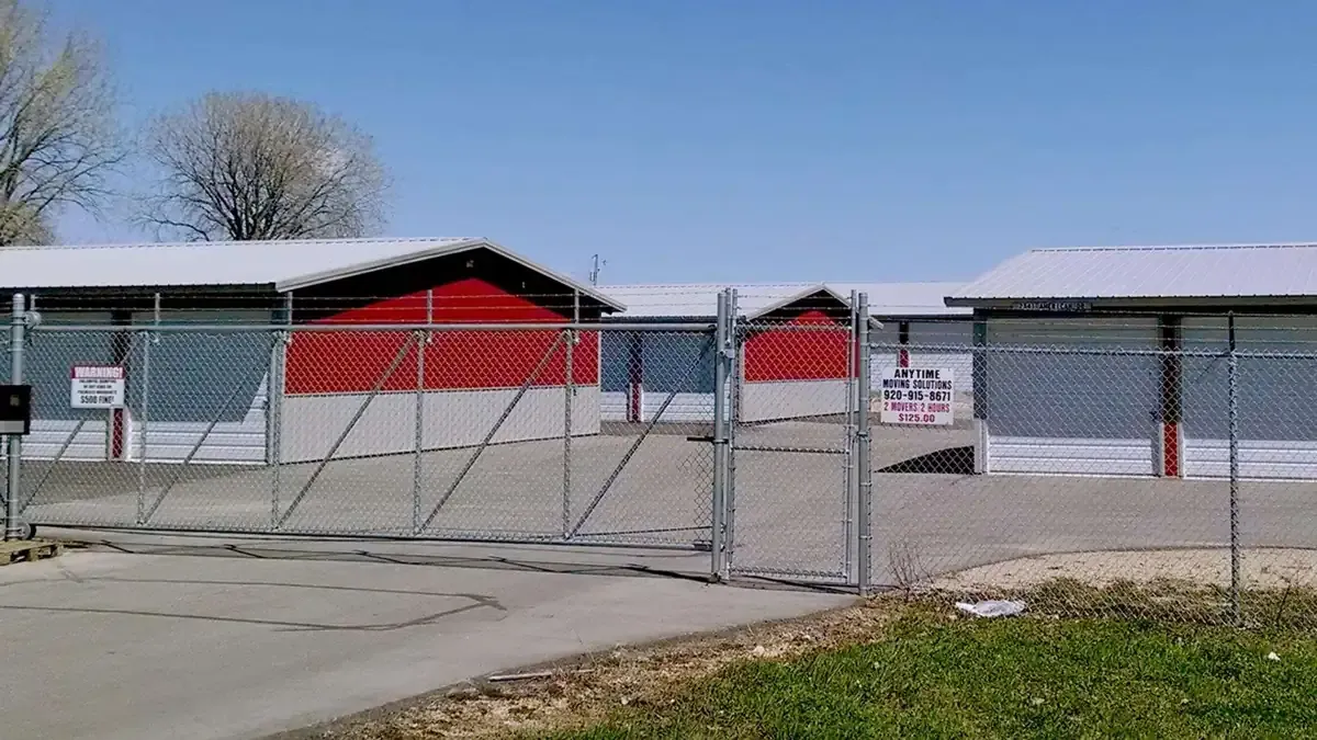A chain-link fence and gate secure a storage facility with red and gray drive-up units.