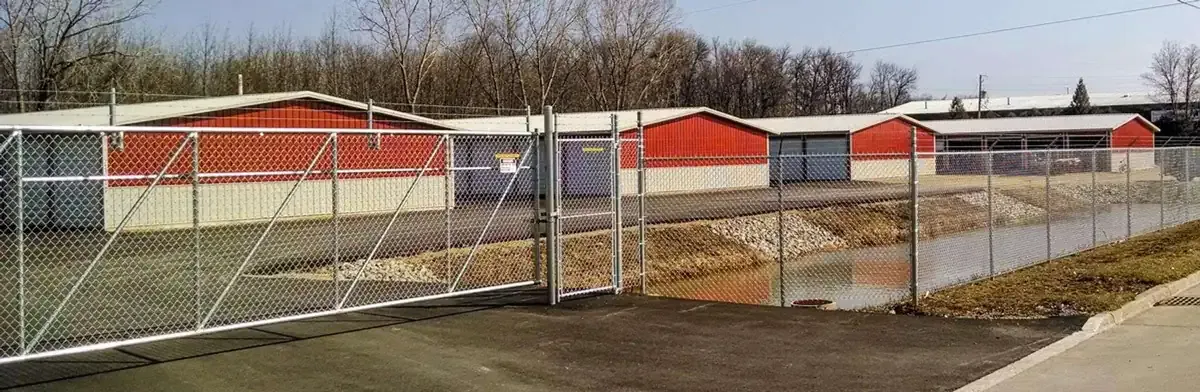 A fenced storage facility with red and white buildings, featuring drive-up units and a chain-link gate.
